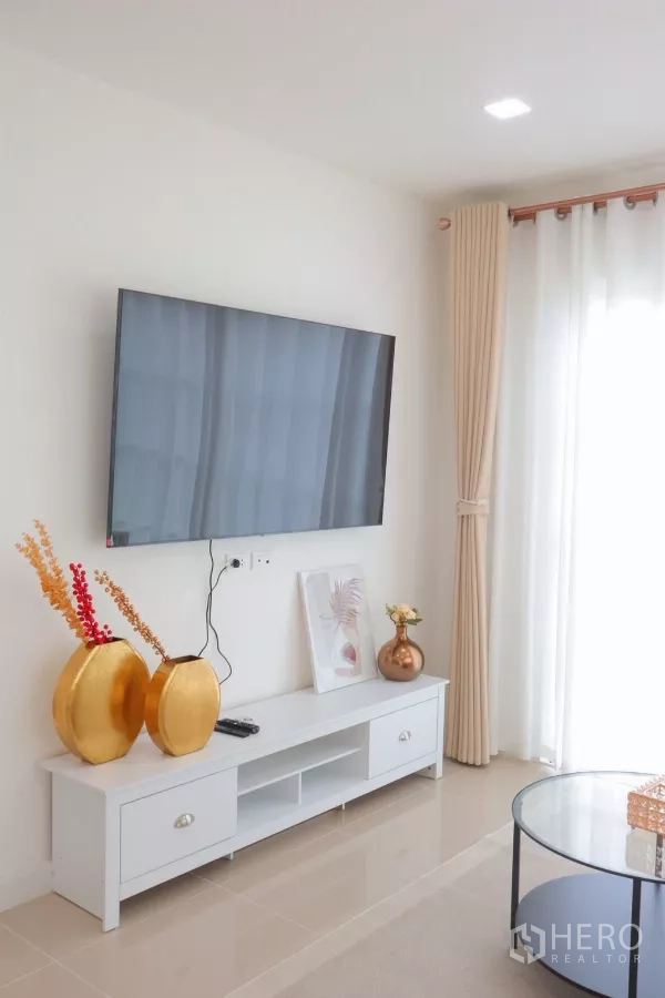 独立式住宅 出租在Bang Phli, Samut Prakan - 电视背景墙，白色矮柜与金色摆件，旁为纱帘落地窗。