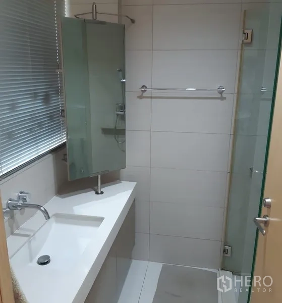 Condominium for rent in Khlong Toei, Bangkok - Contemporary bathroom with long vanity, white basin, window blinds and glass shower enclosure.