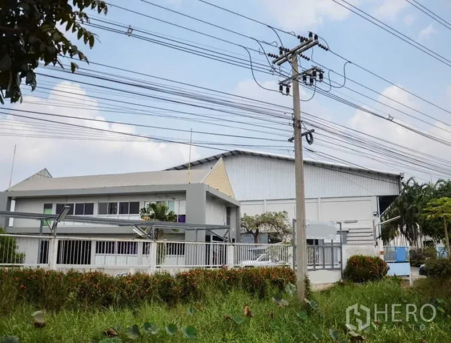 工厂 出租在Pathum Thani - 带围栏的工厂园区，含办公楼与大型仓库，从路边可见电线杆。