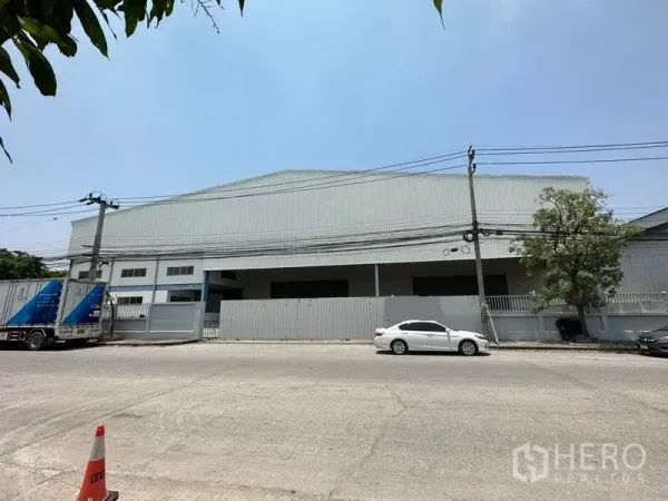 คลังสินค้า ให้เช่าในBang Phli, Samut Prakan - มุมมองด้านหน้าโกดังผนังเมทัลชีท ขอบรั้วติดถนน มีเสาไฟและรถจอด ใต้ท้องฟ้าโปร่ง