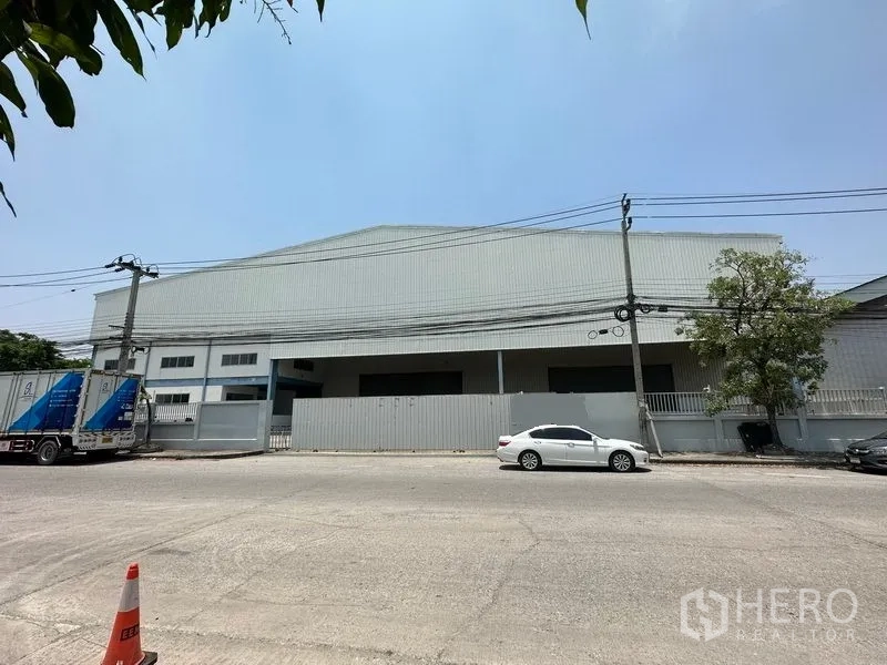 คลังสินค้า ให้เช่าในBang Phli, Samut Prakan - มุมมองด้านหน้าโกดังผนังเมทัลชีท ขอบรั้วติดถนน มีเสาไฟและรถจอด ใต้ท้องฟ้าโปร่ง