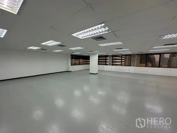 办公空间 出租在Watthana, Bangkok - 开放式办公室，明亮的荧光灯、白色吊顶，窗下长排内置柜体与城市景观。