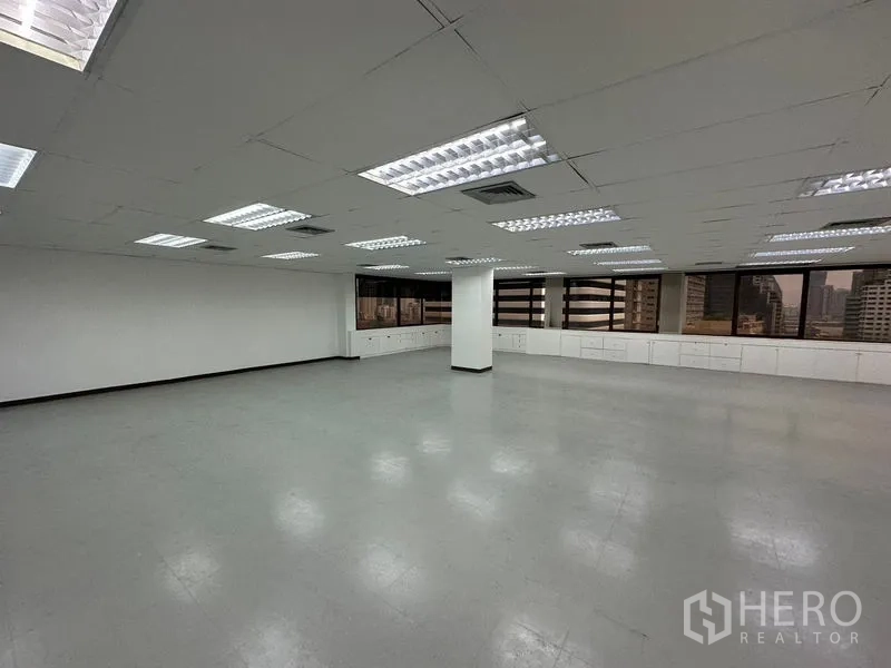 办公空间 出租在Watthana, Bangkok - 开放式办公室，明亮的荧光灯、白色吊顶，窗下长排内置柜体与城市景观。