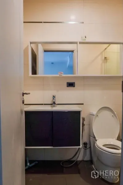 Condominium for rent in Khlong Toei, Bangkok - Compact bathroom with vanity, mirrored cabinet and toilet beside a glass shower.
