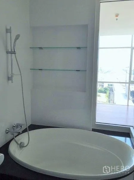 Condominium for sale in Bang Na, Bangkok - Oval soaking tub beside a window with city outlook and built-in glass shelves.