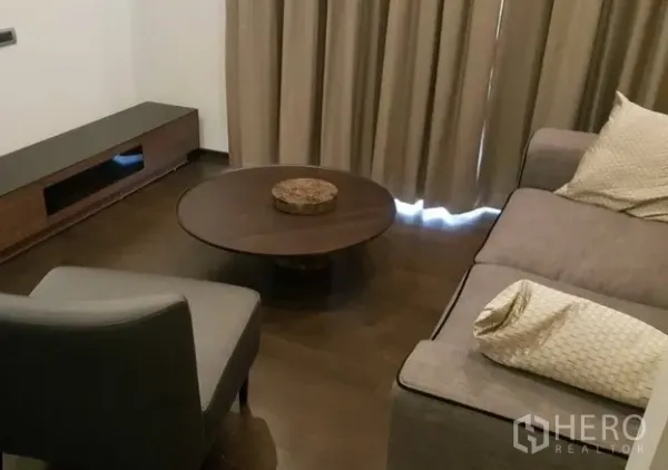公寓 出租或出售在Watthana, Bangkok - 温馨起居区，深色木地板、圆形茶几、电视柜与米色窗帘。