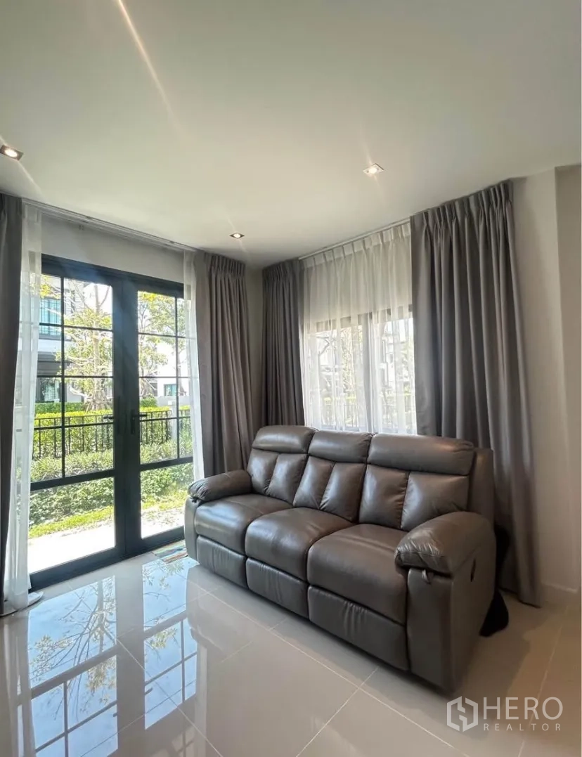Semi-detached House for rent in Bang Phli, Samut Prakan - Cozy living room with gray leather sofa, floor-to-ceiling windows and garden view.