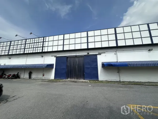 Warehouse for rent in Phra Khanong, Bangkok - Front exterior of white industrial warehouse with blue sliding door, awnings and wide parking apron under a clear sky.