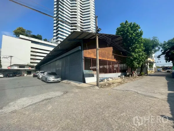 Warehouse for rent or sale in Yan Nawa, Bangkok - Corner view of a single-storey warehouse with metal roof and street access beside high-rise buildings in the Rama 3 area.