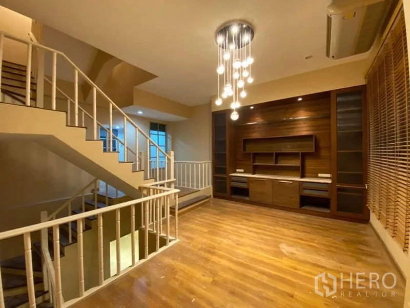 Townhouse for rent in Lat Phrao, Bangkok - Upper family lounge with built-in TV wall, wood floors and statement chandelier beside the stairs.