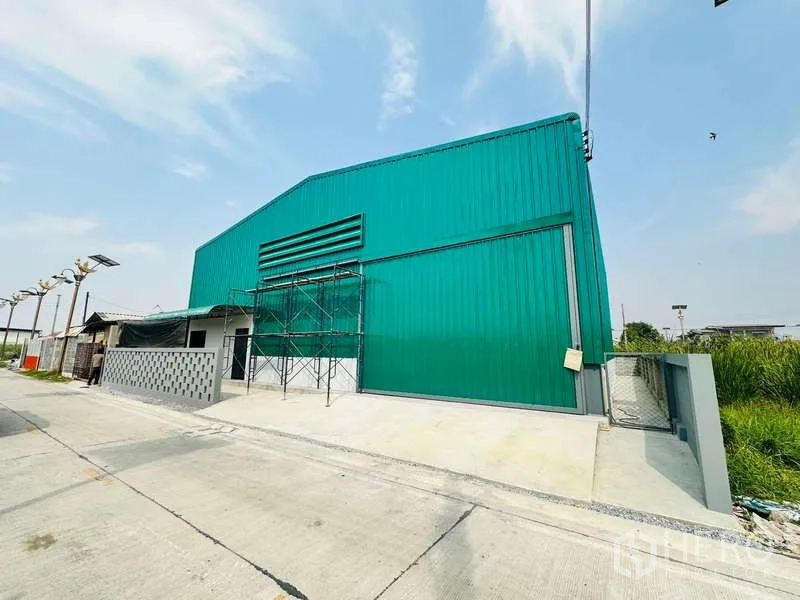 Warehouse for rent in Bang Phli, Samut Prakan - Front facade of green warehouse with large sliding entry door, driveway and gated access for vehicles.