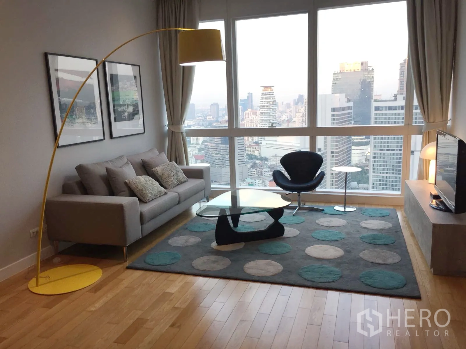 Condominium for rent or sale in Khlong Toei, Bangkok - Bright living room with floor-to-ceiling windows, skyline views, grey sofa, yellow arc lamp and glass coffee table at a Sukhumvit condo.