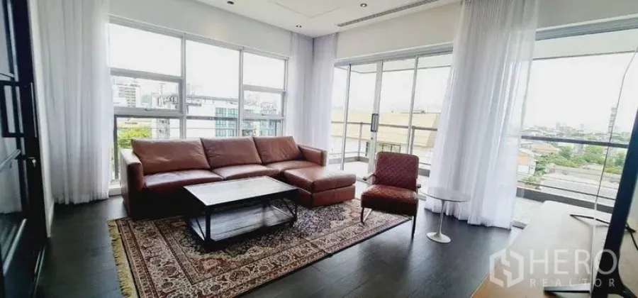 Condominium for rent in Watthana, Bangkok - Corner living room with floor-to-ceiling windows, leather sofa, patterned rug and city views.