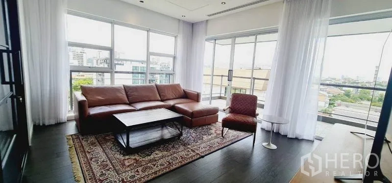 Condominium for rent in Watthana, Bangkok - Corner living room with floor-to-ceiling windows, leather sofa, patterned rug and city views.