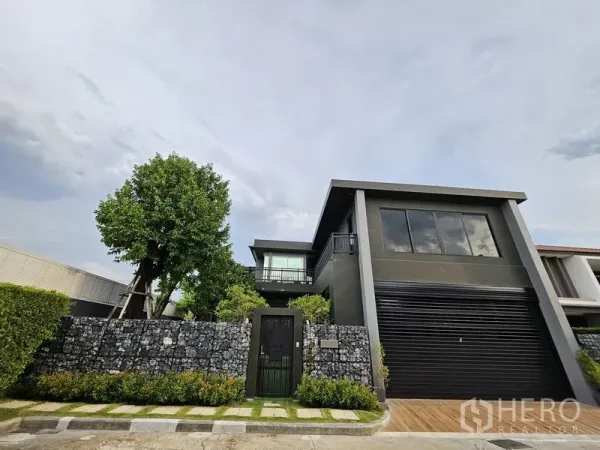独立式住宅 出售在Thawi Watthana, Bangkok - 更广的正面视角，可见石笼围栏与大树。