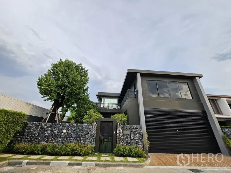 独立式住宅 出售在Thawi Watthana, Bangkok - 更广的正面视角，可见石笼围栏与大树。