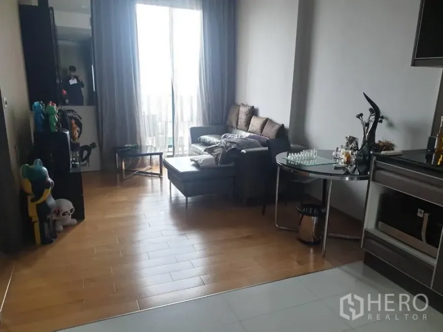 Condominium for rent in Khlong Toei, Bangkok - Bright living room with gray sectional sofa, ottoman, wood floors, and sliding doors to a balcony with sheer curtains.