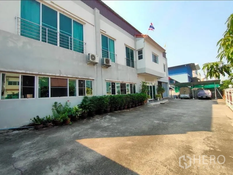 Warehouse for sale in Mueang Samut Sakhon, Samut Sakhon - Side driveway and parking area beside the warehouse with potted plants and a carport.