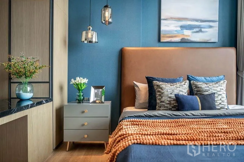 Condominium for rent in Khlong Toei, Bangkok - Bedside table with flowers beneath hanging lamps and upholstered headboard
