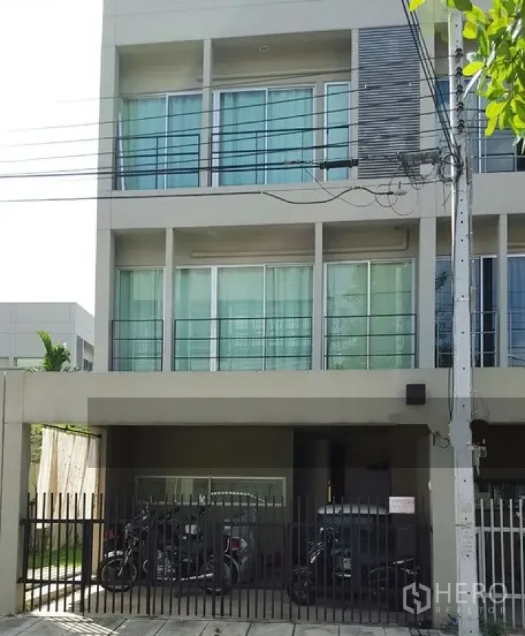 联排别墅 出租在Suan Luang, Bangkok - Noble Cube Suan Luang 现代三层联排别墅外立面，落地推拉窗、纤细阳台，带前院围栏与有顶停车位，环境安静。