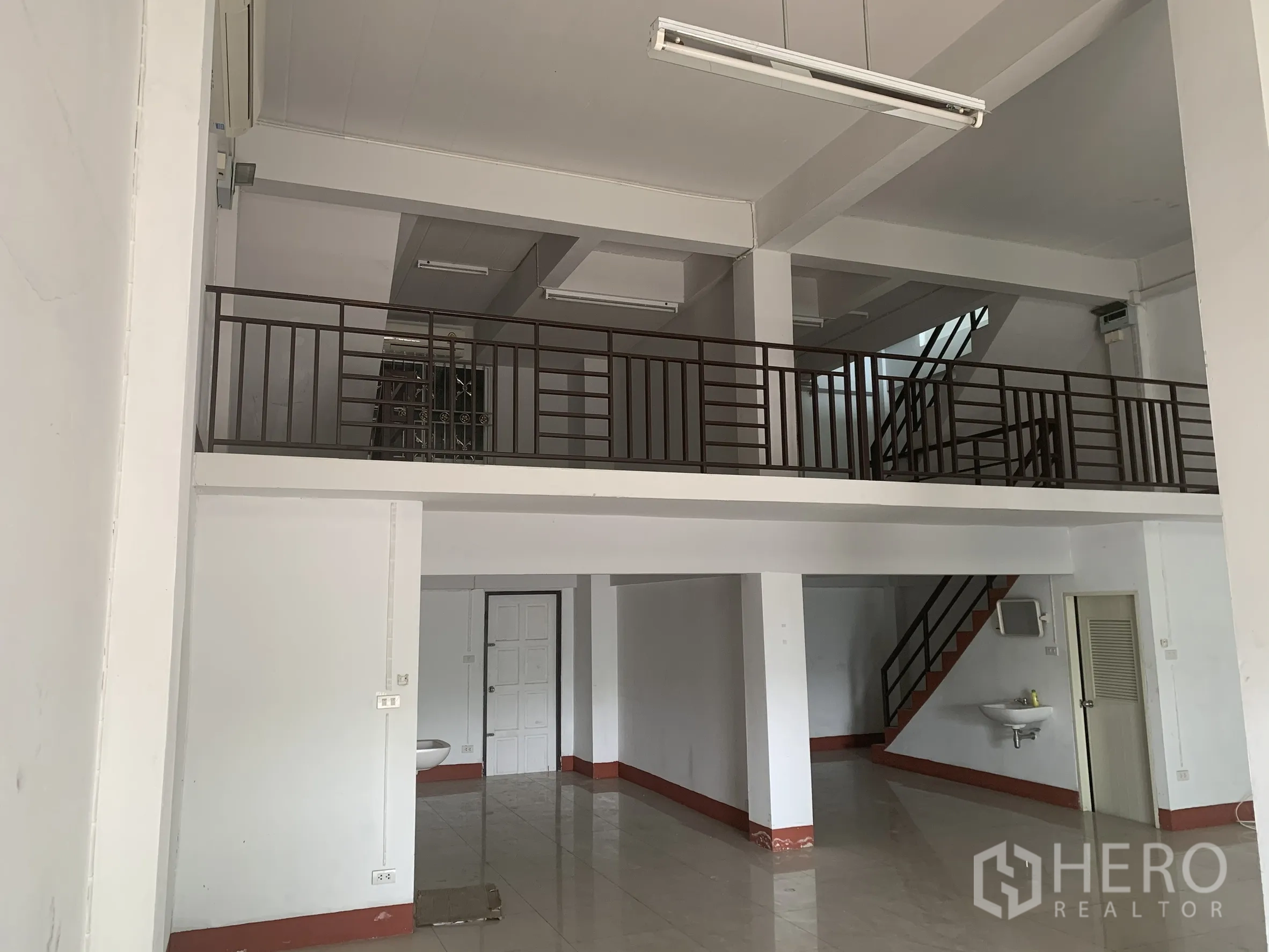 Home Office for rent in Lat Krabang, Bangkok - Double‑height ground floor hall with mezzanine, tiled floors, sink and metal staircase.