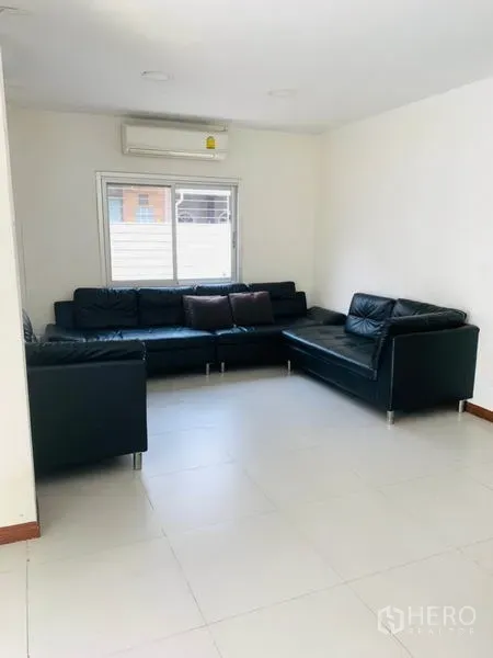 Townhouse for rent or sale in Huai Khwang, Bangkok - Corner lounge with L-shaped black leather sofa, air conditioner and window blinds