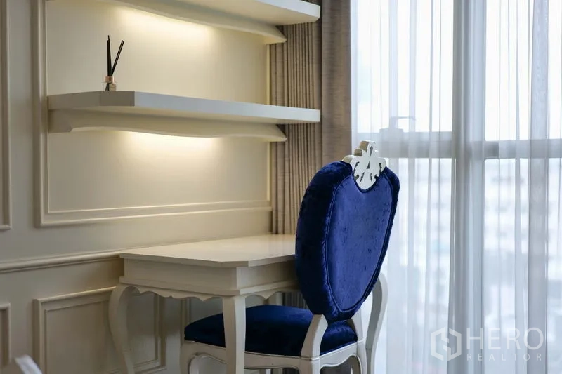Condominium for rent in Khlong Toei, Bangkok - Bright study nook with floating shelves, white desk, and blue velvet chair by the window.