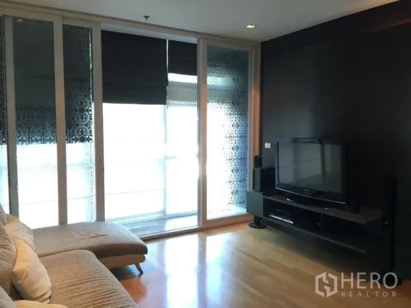 Condominium for rent in Pathum Wan, Bangkok - Cozy lounge with TV console, dark feature wall and sliding glass doors with patterned blinds.