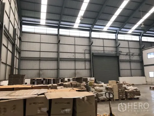 Factory for rent in Khlong Luang, Pathum Thani - Wide warehouse bay with skylights, steel beams, stored pallets and a large roller shutter door.