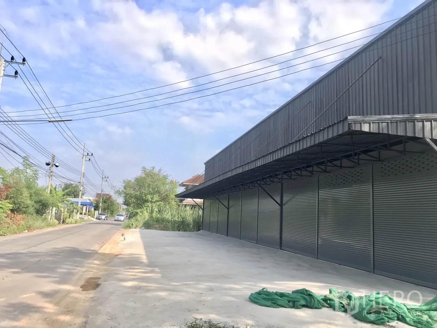Warehouse for rent in Lam Luk Ka, Pathum Thani - Side view of metal-clad warehouse along a local road with power lines and spacious parking apron.