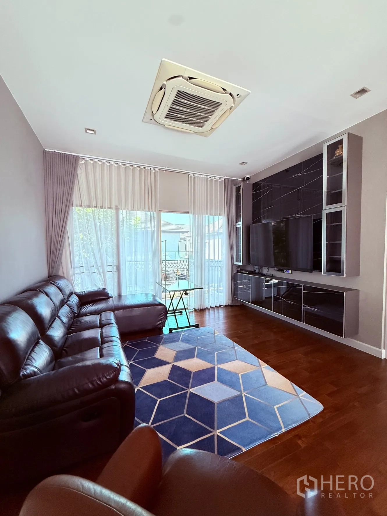Detached House for rent or sale in Prawet, Bangkok - Family room with leather sofa, blue geometric rug, built-in TV wall, and sheer curtains.