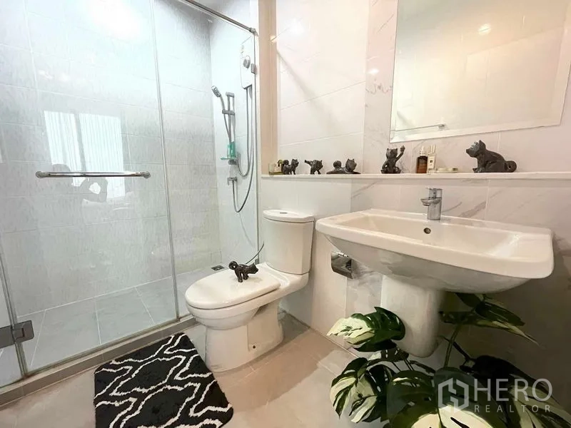 公寓 出售在Huai Khwang, Bangkok - 现代化浴室，玻璃淋浴间、白色瓷砖、立式洗手盆与马桶，并点缀绿植。
