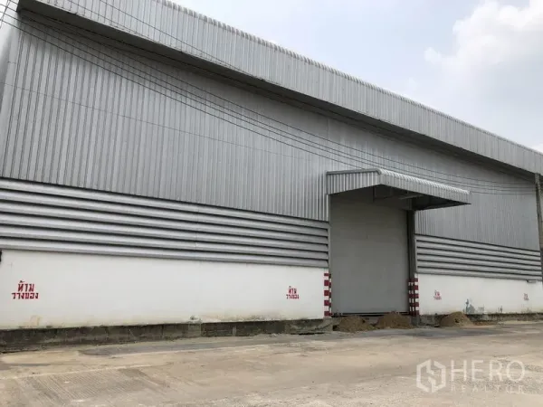 货栈 出租在Pathum Thani - 仓库立面近景，带大卷帘门、金属雨棚与条纹壁板。