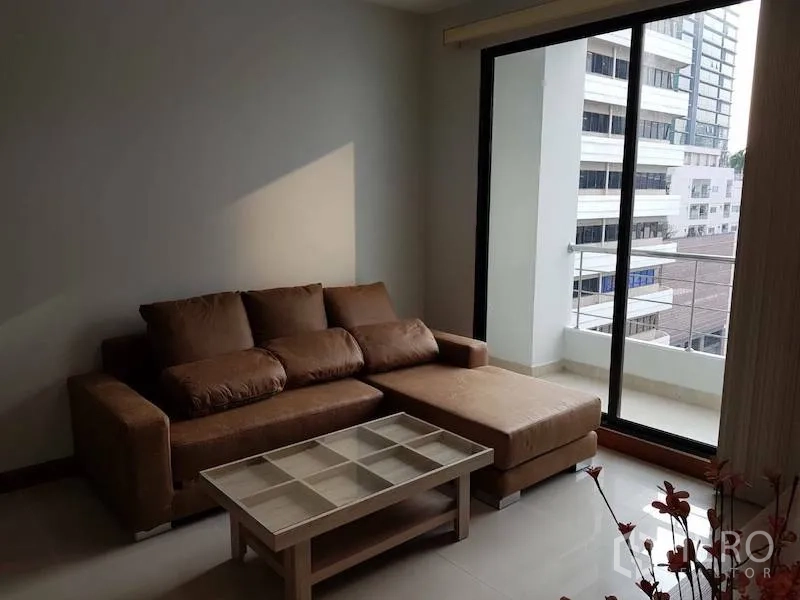 Condominium for rent in Watthana, Bangkok - Living room with brown L-shaped sofa, glass coffee table, and sliding doors to a balcony overlooking city buildings.