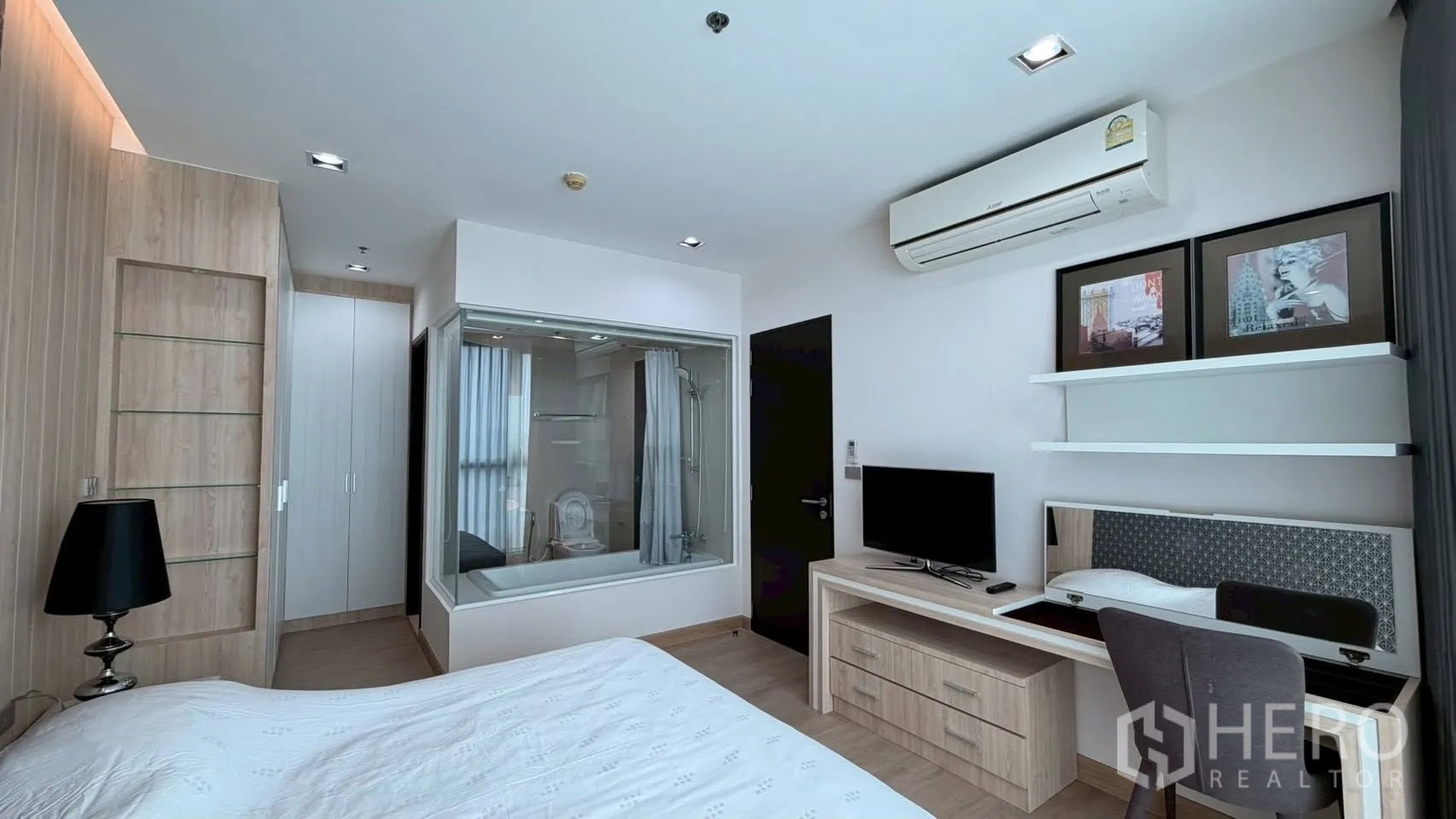 คอนโด for rent or sale in Watthana, Bangkok - มุมห้องนอนมองไปยังห้องน้ำกั้นกระจกที่มีอ่างอาบน้ำและฝักบัว ตู้เสื้อผ้า และโต๊ะทำงานพร้อมทีวี