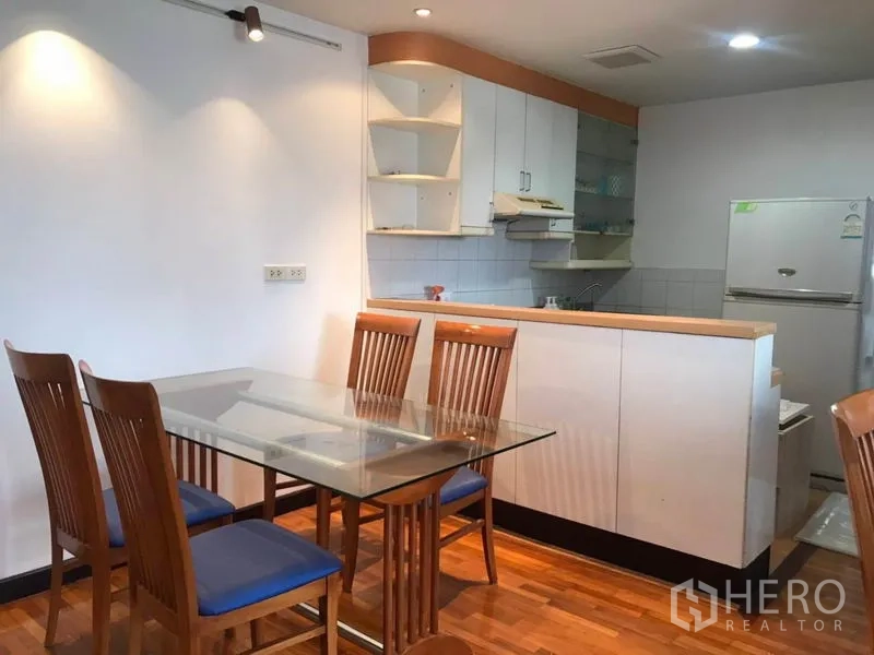 Condominium for rent or sale in Khlong Toei, Bangkok - Glass dining table with wooden chairs beside a compact white kitchen and refrigerator.