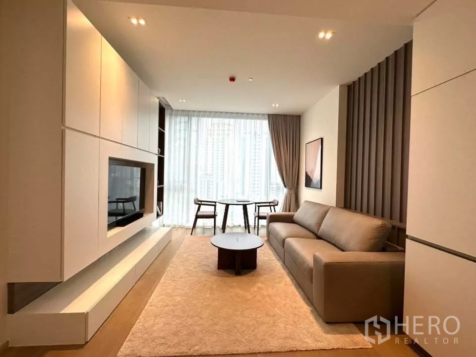 Condominium for rent in Bangkok - Living room with sofa, coffee table, feature wall panels, plush rug, and city views through sheer curtains.