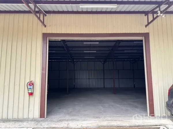 Warehouse for rent in Mueang Nonthaburi, Nonthaburi - Beige metal-clad warehouse facade with wide roller shutter opening, fire extinguisher, and driveway apron.