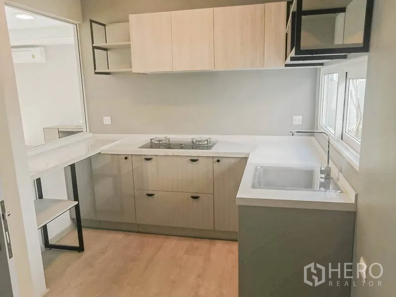 Townhouse for rent in Prawet, Bangkok - Compact L-shaped kitchen with white countertops, gas hob, upper cabinets and a sink by the window.