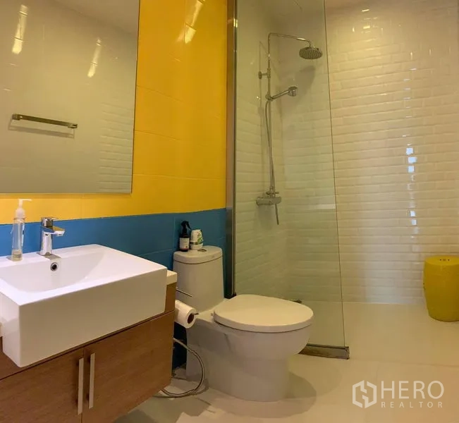 Condominium for rent in Phaya Thai, Bangkok - Modern bathroom with glass-enclosed rain shower, yellow and blue accent tiles, and vanity.