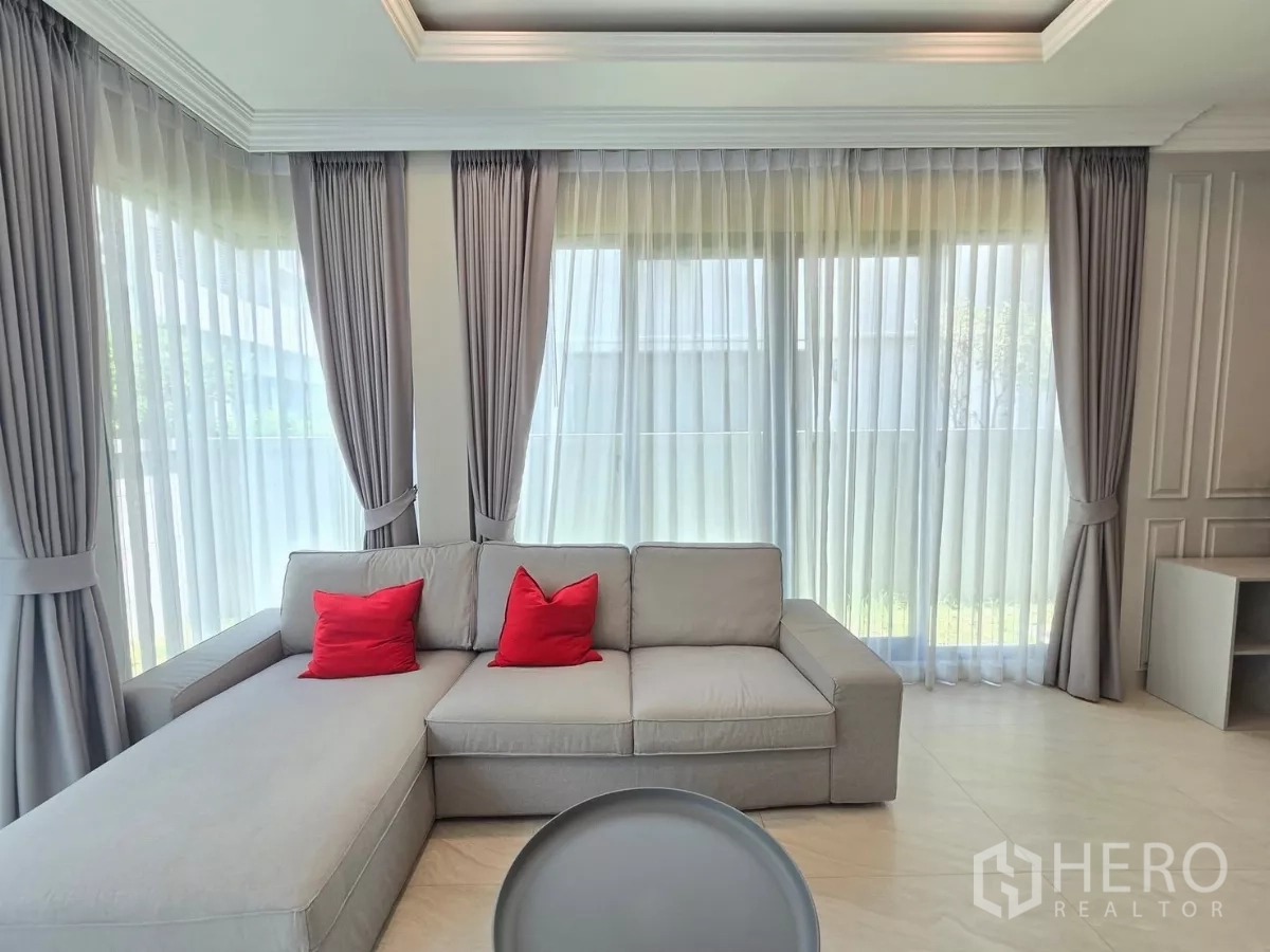 独立式住宅 出租在Prawet, Bangkok - 灰色L型沙发配红色抱枕，旁有高窗与窗帘。