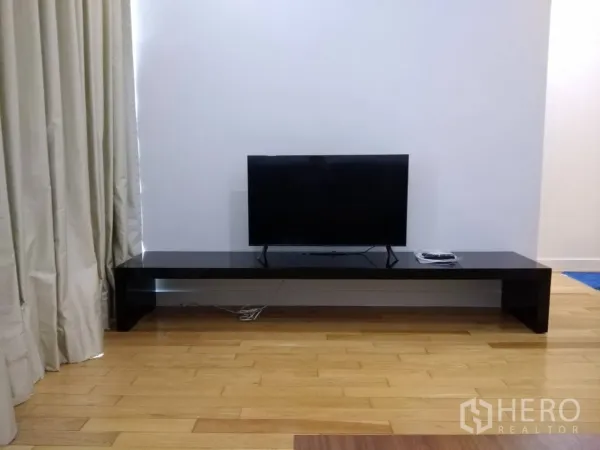 Condominium for rent in Khlong Toei, Bangkok - Flat-screen TV on a low black media console beside tall curtains in the living room.
