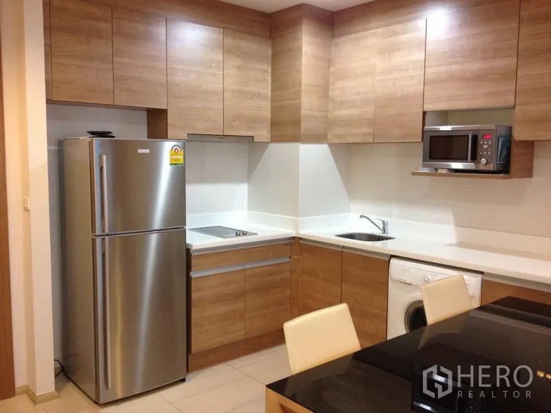 Condominium for rent in Huai Khwang, Bangkok - Modern L-shaped kitchen with wood cabinetry, stainless refrigerator, built-in hob, microwave, sink, and in-unit washing machine.