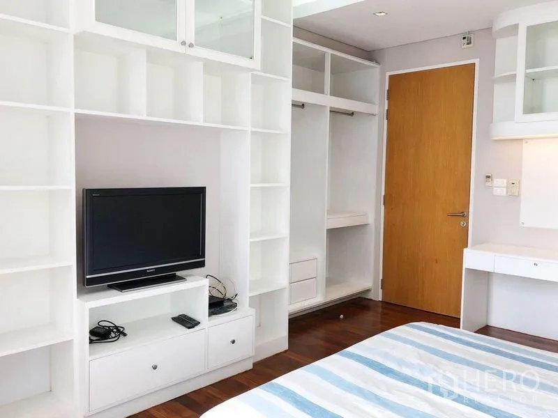 Condominium for rent in Khlong Toei, Bangkok - Bedroom with built-in white shelving, TV unit, open wardrobe and hardwood floors.