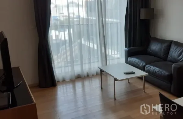 Condominium for rent in Khlong Toei, Bangkok - Bright condo living room with floor-to-ceiling curtains, black leather sofa and coffee table beside a balcony window.