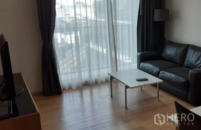 Condominium for rent in Khlong Toei, Bangkok - Bright condo living room with floor-to-ceiling curtains, black leather sofa and coffee table beside a balcony window.
