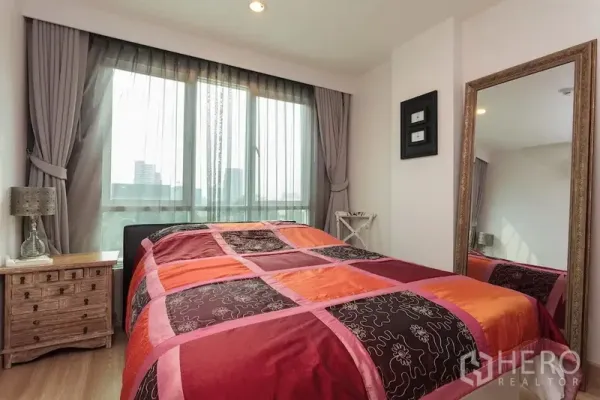 Condominium for rent in Khlong Toei, Bangkok - Sunny bedroom with large window, colorful duvet, bedside table, and full-length mirror.