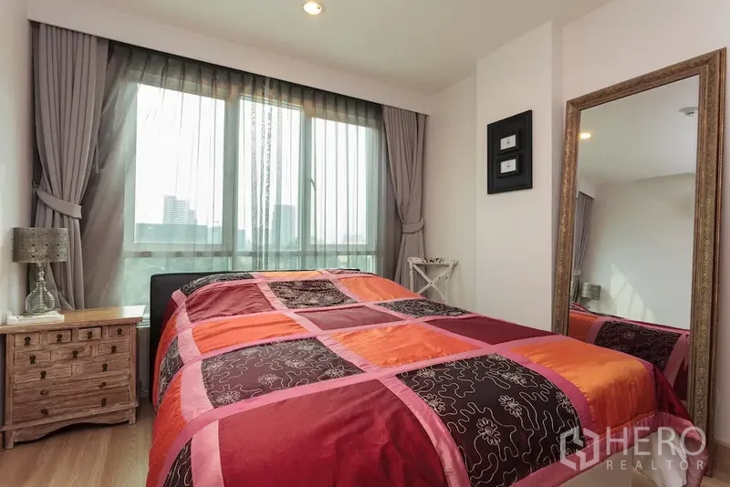 Condominium for rent in Khlong Toei, Bangkok - Sunny bedroom with large window, colorful duvet, bedside table, and full-length mirror.