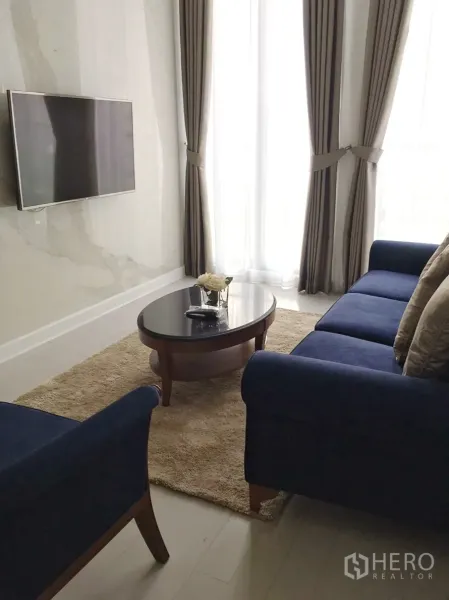 Condominium for rent in Pathum Wan, Bangkok - Cozy lounge area with wall-mounted TV, oval coffee table, and tall draped windows.