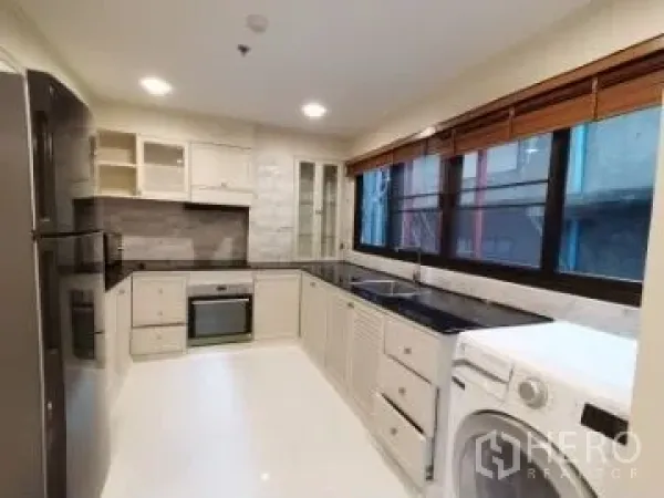 Apartment for rent in Watthana, Bangkok - Modern L-shaped kitchen with white cabinetry, black countertops, oven, fridge, sink, large window and washing machine.